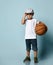 Little child in white t-shirt, gray cap, denim shorts, khaki sneakers. Holding basketball ball, smiling, posing on blue background