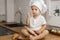 A little cook sits on a table in the kitchen, a 2-year-old boy in a chef`s hat and flour on his cheeks, mom`s assistant