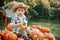 A little cowboy boy in a hat and a plaid shirt is sitting on a pile of pumpkins in the garden and holding a small