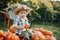 A little cowboy boy in a hat and a plaid shirt is sitting on a pile of pumpkins in the garden and holding a small