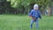 Little cute baby boy in a blue jacket with a toy in hand goes on the lawn to the camera in the city park with trees on