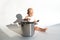 Little cute baby chef sitting near big cooking pot with kitchen, utensils, accessories on white background. Child cook
