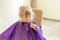 Little cute blond girl smiles and looks at hairdresser during haircut process. Girl holds a wet forelock hair before it will be al