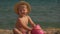 Little cute blond girl in a straw hat plays with sand on the seashore