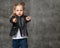 Little cute blond girl in stylish rock style black leather jacket, jeans and sneakers standing and pointing at camera with fingers