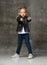 Little cute blond girl in stylish rock style black leather jacket, jeans and sneakers standing and pointing at camera with fingers