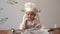 Little cute boy in a chef`s cap sits at a table with flour and eats cookies