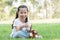 Little cute Caucasian kid girl with twin tails hairs smiling and holding a glass of milk and sitting on green grass at park