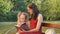 Little Cute Daughter with Two Ponytails Reading Book with Her Mother in Eyeglasses Sitting on the Bench in the Park.