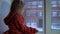Little cute girl sitting on the window sill, looking out on a snowy cityscape.