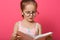 Little cute girl wearing summer dress and glasses, holds book in hands, looks concentrated, reading interesting fairy tale, poems