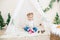 Little cute girl in a white warm knitted cardigan near a children`s lodge decorated with a garland and needles, toys