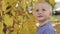 Little cute one year old baby is played near the tree in the autumn park. Standing on fallen yellow leaves and smiling