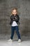 Little cute smiling blond girl in stylish rock style black leather jacket, jeans and white sneakers standing in confident pose