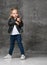 Little cute smiling blond girl in stylish rock style black leather jacket, jeans and white sneakers standing in confident pose