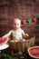 Little cute toddler with blond hair sits in basket with watermelon on a dark wooden background. Children and fruits.