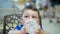 Little dark-eyed boy is sitting on a chair and drinking water from baby bottle in cafe, close up