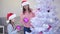 Little daughter holds a lot of pink glass baubles, her mother takes baubles and decorates a fir-tree.