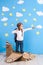 Little dreamer girl playing with a cardboard airplane at the studio with blue sky and white clouds background.