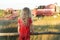 Little farm girl wearing red polka dot kids pans looking at field with working combine harvesters
