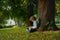 The little fellow sits under a big tree and looks at the tablet screen.