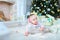 Little female baby lying on floor near twinkling Christmas tree and presents.