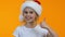 Little female kid in santa hat showing thumbs-up and winking on camera, holiday