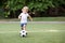 Little football player: blonde child in white shirt and blue shorts running along the green soccer field ready to kick ball.