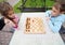 Little frowning girl and smiling boy play chess at table in park