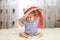 A little funny caucasian little girl a child sits at home on the floor on a light carpet. On his head is a large summer hat thatch