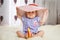 A little funny caucasian little girl a child sits at home on the floor on a light carpet. On his head is a large summer hat thatch