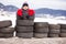 Little funny child in red jacket standing inside used tires among the road mountain view on background