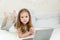 A little girl in bed with a laptop. A 4-year-old girl with long loose hair in a white T-shirt on a white bed looks directly into