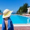 Little girl with a blue towel and a straw hat stands with her back and looks at the pool