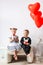 Little girl and boy sitting on a white chair near heart-shaped baloons. Girl licking a red lollipop. Valentines day