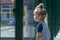 A little girl carefully looks over the fence at the children`s playground. Concept: the desire to play sports, the limitations of