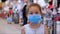Little girl child in a mask from viruses of the epidemic of the coronavirus or viruses looks at the camera among people
