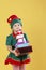 Little girl in a Christmas elf costume, carries a stack of gifts. Photo on a yellow background.