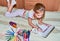 A little girl draws in the album with colored pencils  in a room on a bed  next to lie colored pencils  scissors  toys. Side view