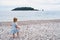 Little girl in a dress walks along a pebble beach to the sea with an island in the distance