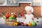 Little girl dressed in white chef hat and apron, sits among vegetables and holds an apple.