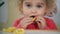 Little girl eating corn circle sitting near table wit toys and drawings on wall