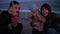 Little girl on father`s arms watching her mother, father waving sparklers on the beach in the evening dusk. Happy young