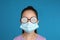 Little girl with foggy glasses caused by wearing medical face mask on blue background. Protective measure during coronavirus