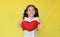 Little girl giving red heart for Valentine`s Day for you on yellow background. Focus at kid face