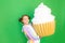 A little girl on a green isolated background in a bright suit holds a huge cake. Space for text. Concept of junk food and weight