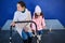 Little girl with her mother pushing baby stroller, sitting on the banc at railway station platform, waiting for train