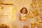 Little Girl Holding Candle sitting next to Christmas Tree Decorated with Golden Ornaments and Lights. Happy Child in White Room