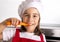 Little girl at home kitchen in red apron and cook hat holding carrot and biting happy