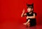 Little girl with horns, in black blouse and leggings. Looking at her raised forefinger, sitting on floor, red background.